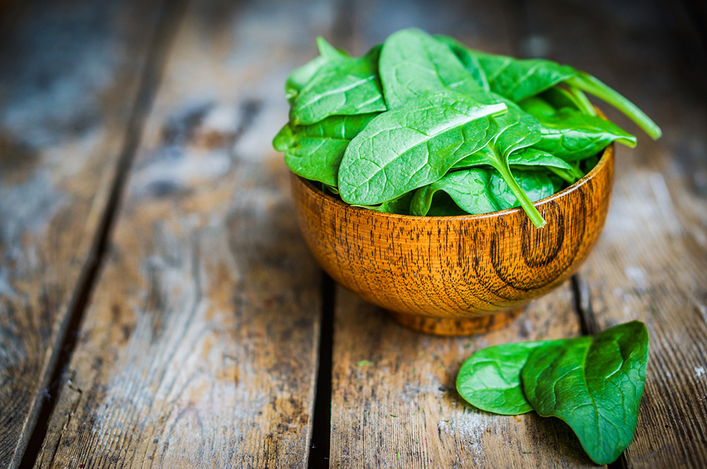 A wooden bowl of spinach sitting next to some spinach leaves on a wooden table.